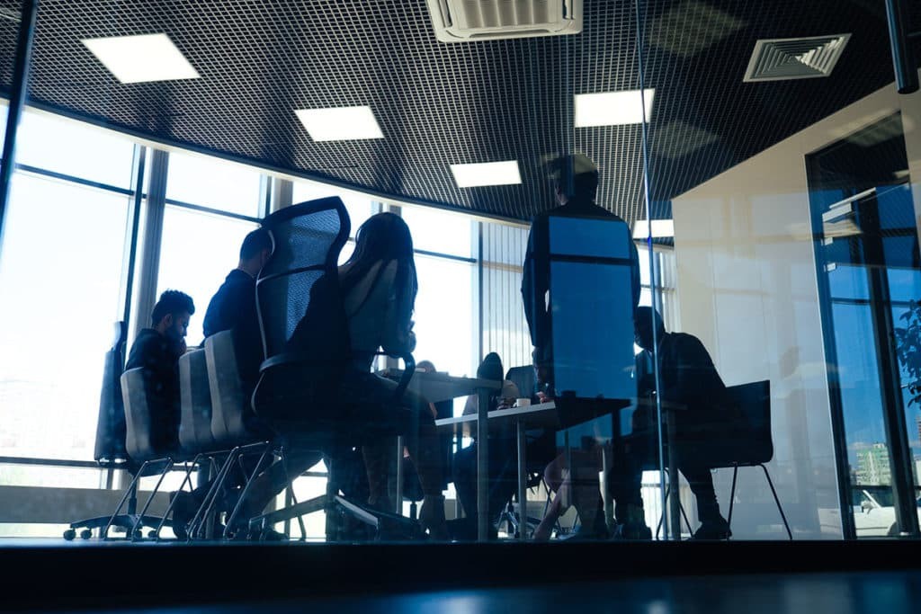 Meeting room with glass walls, seen from outside, with people around the table discussing and taking notes.